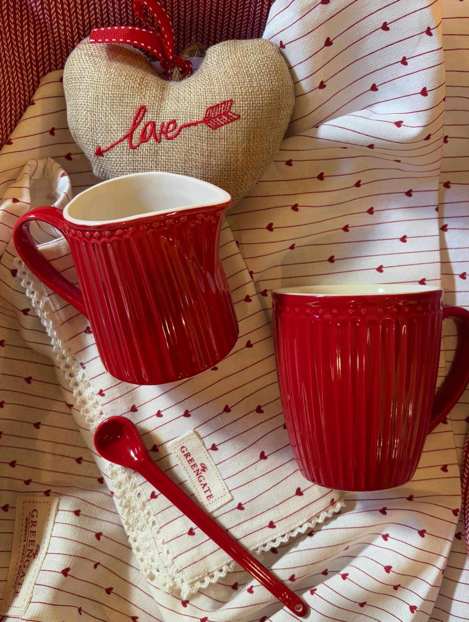Bodegón de estilo romántico con una jarrita y una taza de cerámica roja con relieve de líneas verticales de la marca GreenGate. Al fondo, un corazón de tela de saco con la palabra "love" bordada y, debajo, una cuchara de cerámica roja sobre un paño de cocina blanco con pequeños corazones rojos.