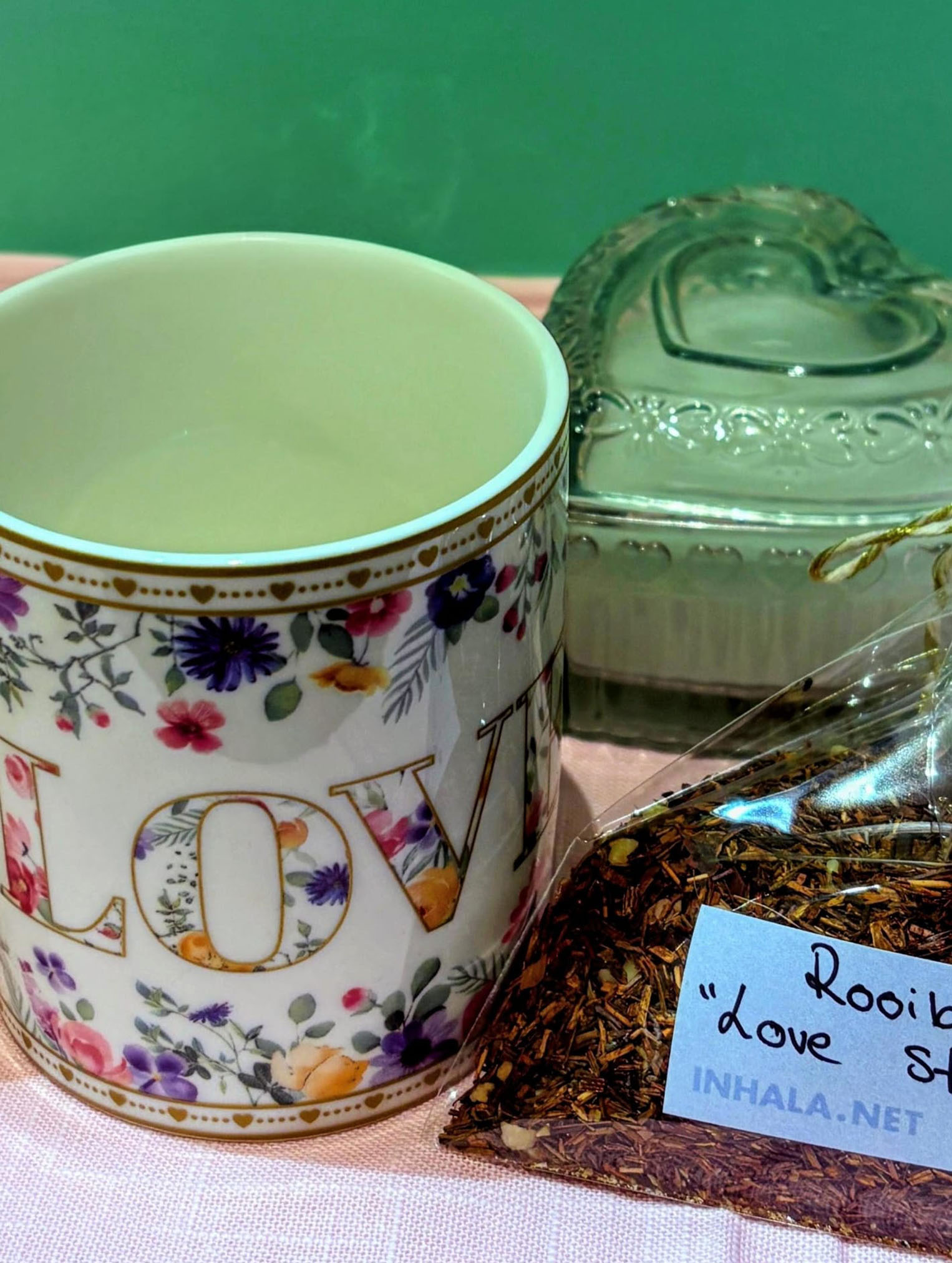 Primer plano de una taza blanca decorada con flores coloridas y la palabra "LOVE". A su lado, un paquete de té rooibos etiquetado como "Love Story" de Inhala y, al fondo, una vela artesanal dentro de un recipiente de cristal con forma de corazón.