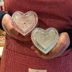 Una persona con un delantal de rayas rojas y blancas de la marca GreenGate sostiene con ambas manos dos recipientes de cristal con forma de corazón y tapas ornamentadas. Uno de ellos contiene una vela con la etiqueta "Magnolia Vanilla". Regalar-se un moment bonic de cafès, tes i infusions és un regal per un mateix. Al final, l'Amor propi és el que més val. Triar un cafè especial i preparar-lo disfrutant de les seves aromes. I per què no? Acompanyar-lo d'una xocolata o galeta, petits moments de felicitat. Així, doncs, avui un pensament especial per l'AMOR.            INHALA Cafès i Tes                 Portalet, 9           08401 Granollers 