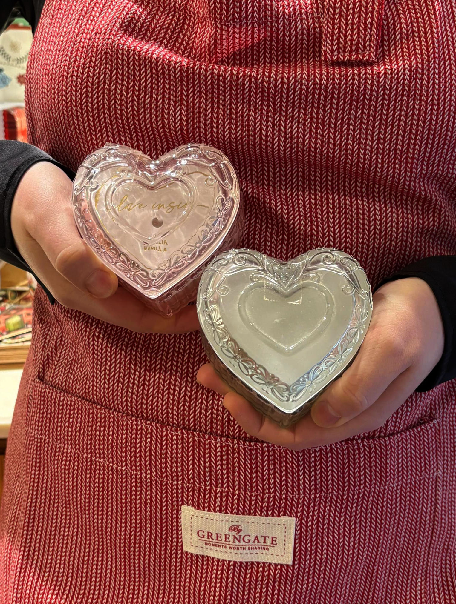 Una persona con un delantal de rayas rojas y blancas de la marca GreenGate sostiene con ambas manos dos recipientes de cristal con forma de corazón y tapas ornamentadas. Uno de ellos contiene una vela con la etiqueta "Magnolia Vanilla".