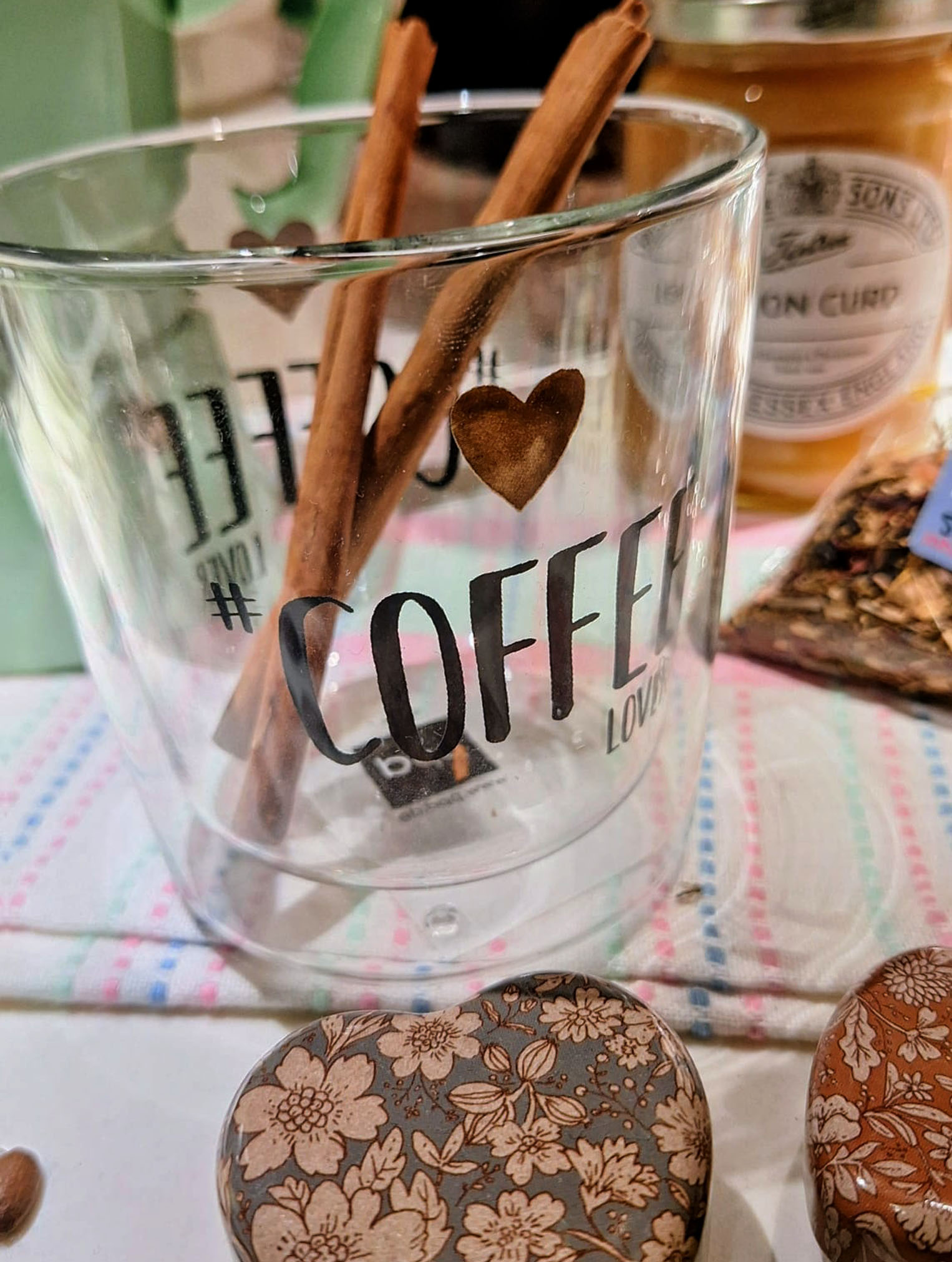 Un vaso de cristal de doble pared con el texto "#COFFEE LOVER" y un corazón, que contiene dos ramas de canela. En primer plano se ven cajas metálicas con estampados florales y, al fondo, un tarro de "Lemon Curd" y una bolsa de té.